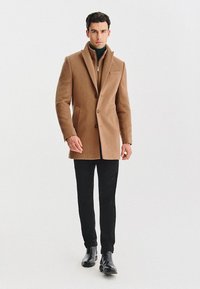 Brązowy płaszcz z mieszanki wełny o dopasowanym kroju, z klapami, jednolitym zapięciem na guzik, bocznymi kieszeniami, połączony z golfem i czarnymi spodniami.