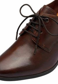 Chaussure en cuir marron avec une surface lisse, bout arrondi, design à lacets, et lacets marron foncé. Présente des œillets en métal et des détails de couture.