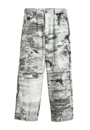 Joven con rastas que lleva chaqueta y pantalones grises con un patrón abstracto, camisa azul marino, zapatos marrones y una larga correa con estampado que cuelga de la cintura.