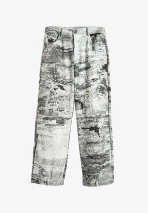 Joven con rastas que lleva chaqueta y pantalones grises con un patrón abstracto, camisa azul marino, zapatos marrones y una larga correa con estampado que cuelga de la cintura.