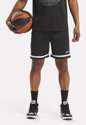 Schwarze Sportshorts mit weißen Seitstreifen, Netzstruktur, kombiniert mit schwarzen Turnschuhen und einem Basketball. Modell steht und hält den Ball.