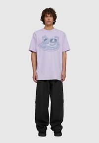 Fialové tričko s modrou grafikou, která obsahuje čísla a hvězdy, kombinované s volnými černými kalhotami a černými botami. Hladká textura, oversized střih.