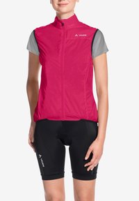 Gilet antivento fucsia con maniche corte grigio tenue, cerniera frontale e logo riflettente, abbinato a pantaloncini da ciclismo neri.