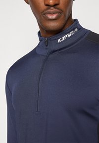 Laivyno mėlynos zip-up ilgos rankovės palaidinė su aukštu apykakle, pasižyminti sklandžiu paviršiumi ir logotipu ant kaklo. Šoninės dalys iš kontrastingos medžiagos.