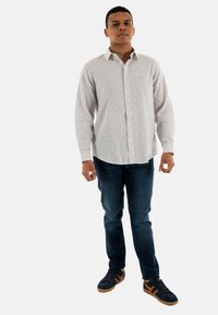 Camicia bianca a maniche lunghe con bottoni e sottili righe, abbinata a jeans blu e sneaker nere con dettagli marroni.