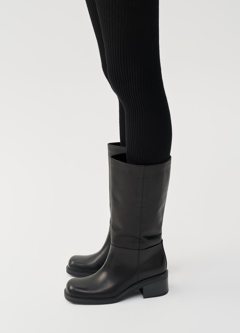Persona che indossa leggings neri a costine e stivali in pelle nera fino a metà polpaccio con tacco basso a blocco, in piedi su uno sfondo chiaro e uniforme.
