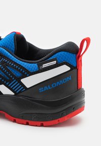 Zapatilla de trail negra y azul con acentos rojos, que cuenta con una suela texturizada, etiqueta impermeable y un diseño de franjas blancas distintivas.