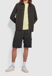Chaqueta ligera gris oscuro con dos bolsillos en el pecho, combinada con una camiseta a rayas amarillas y blancas y pantalones cortos de cargo negros. Zapatillas blancas con detalles verdes.