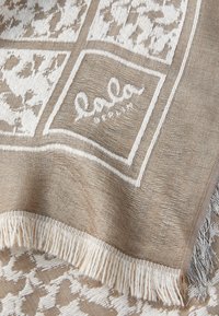 Beige vevd teppe med hvite geometriske mønstre og frynsete kanter. Har et logo brodert i hvitt, som viser et teksturert stoffdesign.