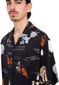 Camisa de manga corta, con botones, en tela negra que presenta ilustraciones de animales, mariposas naranjas y texto en blanco. Bolsillo en el pecho izquierdo.