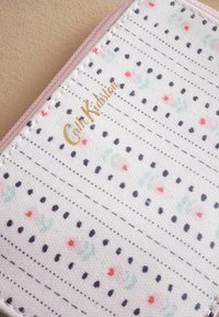 Gepubliceerd stoffen portemonnee met een witte achtergrond, voorzien van zwarte stippen, pastelkleurige bloemenaccenten en een gouden "Cath Kidston" logo langs de rand.