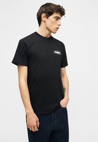 Νεαρός άνδρας με κοντά σγουρά μαλλιά που φοράει μαύρο μπλουζάκι με μικρό λευκό λογότυπο «OBEY» στο στήθος, τα χέρια στις τσέπες σκούρων μπλε τζιν.