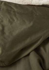 Olijfgroene buitenlaag met een gladde textuur, voorzien van een beige fleece voering aan de bovenrand, zichtbaar stiksel.