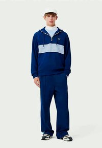 Dunkelblaues Sweatshirt mit einem hellblauen horizontalen Streifen, auf dem "LACOSTE" geprägt ist. Kombiniert mit passenden dunkelblauen Hosen und grauen Sneakers.