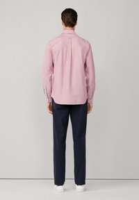 Hombre de pie de espaldas, vestido con una camiseta de manga larga rosa, pantalones oscuros y zapatillas blancas, contra un fondo claro y liso.