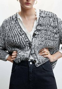 Schwarz-weiße Strickjacke mit strukturiertem Muster, tiefem V-Ausschnitt und großen Knöpfen, getragen über einem weißen Shirt mit Rüschendetails.