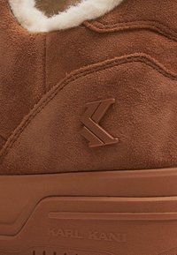Pruun suede spordisaabas, millel on kohev valge vooder, tekstureeritud kummitald ja pruuni logoga accent külgedel.
