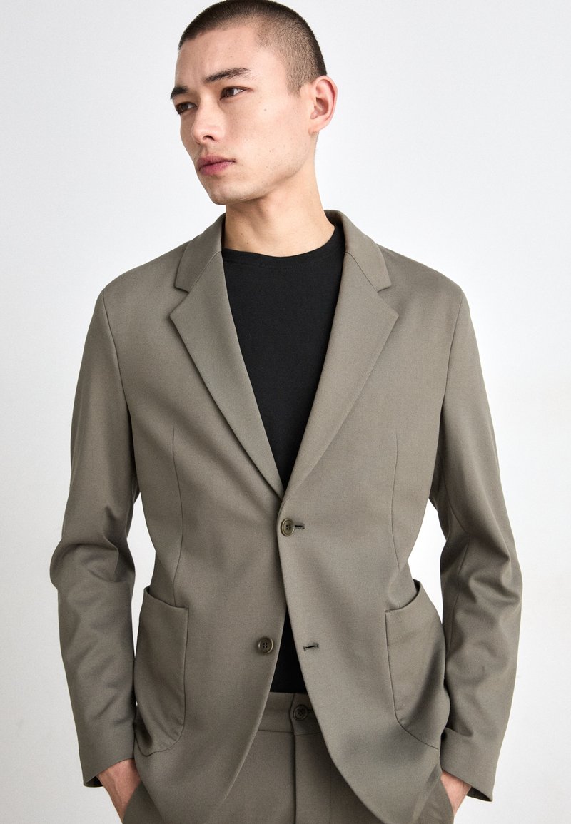 Olivengrønn blazer med skrå lapel, to frontlommer og én knappelukking, laget av glatt, strukturert stoff.