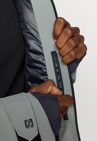 Kezek egy szürke dzseki belső zsebének felhúzásán dolgoznak, amely egy fekete, textúrázott ujjú felső felett viselt.