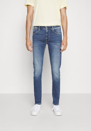 Homme portant un jean slim bleu avec des détails délavés et des baskets blanches, debout devant un fond clair uni.
