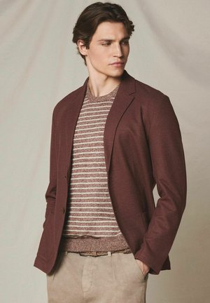 Junger Mann trägt ein braunes Sakko über einem gestreiften beige-braunen Pullover mit beigen Hosen, schaut seitlich und hat die Hände in den Taschen.