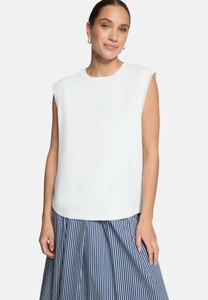 Femme aux cheveux tirés en arrière portant un haut blanc sans manches avec des bords festonnés et une jupe à rayures verticales bleues et blanches.