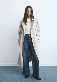 Beige dubbelzijdig trenchcoat over een wit geribd topje, met donkerblauwe wijde jeans en zwarte schoenen op een lichtblauwe tapijt.