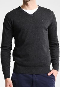 Pull en V en tricot gris foncé avec manches longues, poignets ajustés et un petit logo sur la poitrine. Design simple et classique.