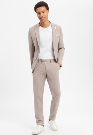 Junger Mann in beigem Anzug mit weißem T-Shirt und weißen Turnschuhen, steht mit einer Hand in der Tasche und lächelt, vor einfarbigem Hintergrund.