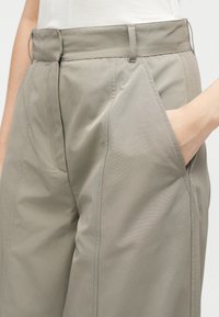 Κοντινό πλάνο μπεζ παντελονιού με ψηλή μέση, πλαϊνές τσέπες, φερμουάρ μπροστά και ένα χέρι που βρίσκεται μέσα στην δεξιά τσέπη.