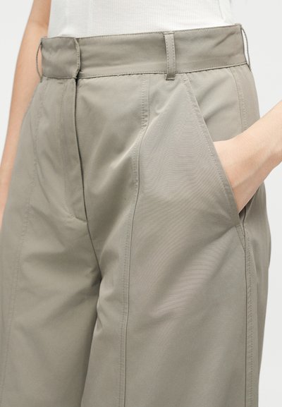 Gros plan sur un pantalon beige taille haute avec poches latérales, fermeture éclair à l'avant, et une main placée dans la poche droite.