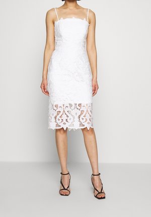 Robe blanche ajustée avec des détails complexes en dentelle, un ourlet festonné et des bretelles fines, accompagnée de talons noirs à lanières.