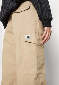 Pantalon cargo Carhartt beige avec poches à rabat boutonnées, porté avec un haut à manches longues rayé noir et gris foncé, et une main visible.
