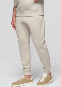 Homme portant un pantalon de jogging beige et des baskets en daim beige assorties, debout avec la main gauche dans la poche du sweat-shirt.