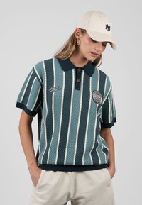 Mujer que lleva una camisa polo de punto rayada azul verdosa, gorra beige con logo y pantalones claros, mirando hacia abajo con las manos en los bolsillos.