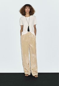 Beige fluwelen wijde broek met een zachte textuur, gecombineerd met een eenvoudige witte t-shirt en een lange zwarte kwastketting. Eenvoudige sandalen maken de look compleet.