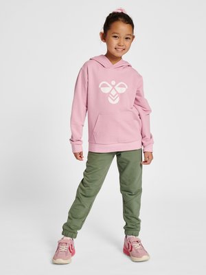 Lächelndes junges Mädchen, das einen pinken Hoodie mit weißem Logo, grüne Jogginghosen und pinke Sneakers trägt, mit gespreizten Beinen auf weißem Hintergrund steht.
