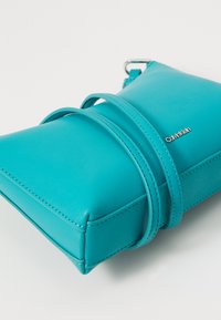 Turkos läder crossbody-väska med rektangulär form, har justerbara axelremmar och en silverfärgad logotypaccent på sidan.