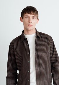 Braun gestreiftes Button-Down-Hemd über einem weißen Rundhalsausschnitt-T-Shirt; hat eine Brusttasche und blaue Knöpfe, glatte Textur, moderne Passform.
