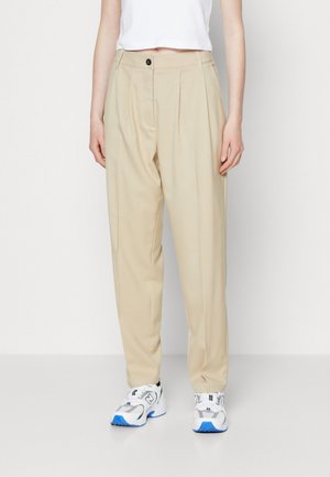 Pantaloni bej cu talie înaltă, cu pliuri și închidere frontală cu buton, asortați cu adidași albi și bleumarin. Textură de țesătură fină.