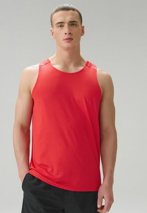 Noor mees lühikeste juustega, seljas punane varrukateta spordisärk ja mustad lühikesed püksid, seisab ühevärvilise halli tausta ees.