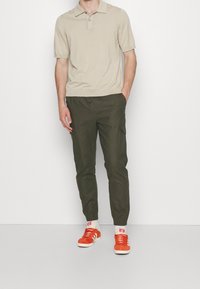 Beige Poloshirt, olivgrüne Cargo-Hose und rote Sneakers mit weißen Sohlen. Lässiger Schnitt, kurzärmlig und leichte Textur.