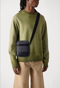 Black shoulder bag with horizontal zipper pockets, made of fabric and leather. Worn on a green sweatshirt against a light background.