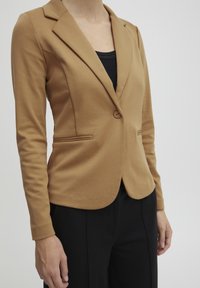 Blazer marrón ajustado con un botón, mangas largas y bolsillos laterales. Tejido suave con una silueta ajustada. Lleva puesto un top negro.