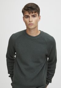 Pulover tricotat de culoare verde închis, cu o textură ribbată, decolteu rotund și mâneci raglan. Design simplu, urmează un stil clasic.