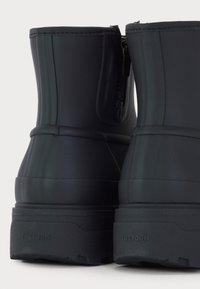 Stivaletti neri impermeabili alla caviglia con suola in gomma testurizzata, dettaglio con zip sul lato e finitura liscia. Logo Tretorn visibile sul tallone.