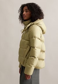 Hellgrüne Pufferjacke mit Kapuze, gestepptem Design, Frontreißverschluss und Kordelzug. Über einem karierten Rock getragen.