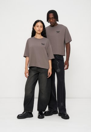 To modeller bærer løstsiddende grå T-shirts med sommerfugle-logoer, kombineret med brede sorte jeans og sorte kraftige sko mod en ensartet hvid baggrund.