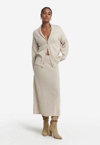 Beige stickad kofta med sjalkrage, framknappar och ribbade detaljer, tillsammans med en lång, matchande kabelstickad kjol och klackboots.