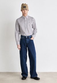 Karierte Bluse mit Knopfleiste in Rot, Blau und Weiß; kombiniert mit lässigen blauen Denim-Jeans; khakifarbene Mütze; schwarze Schuhe.
